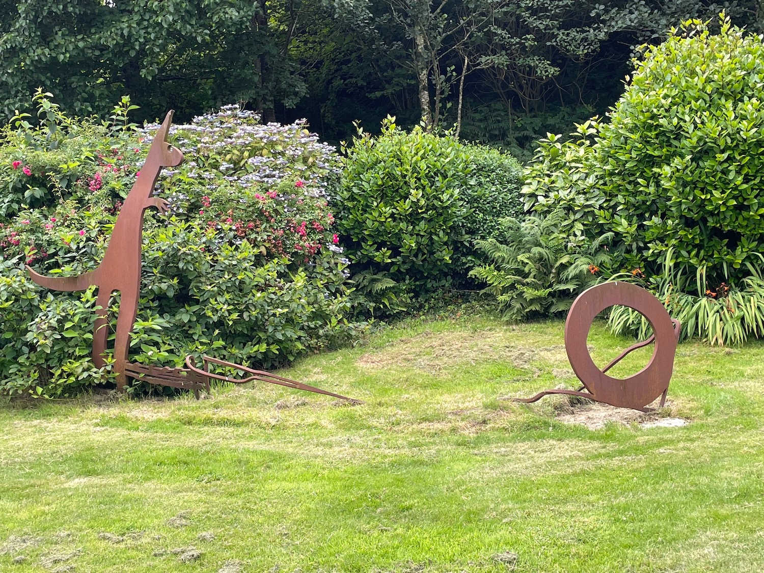 Chasing the Visual Element – sculpture in metal (rusted surface) in a garden setting; to the left is the profile of a cartoonish kangaroo, to the right a circular shape in metal which the kangaroo may be pursuing